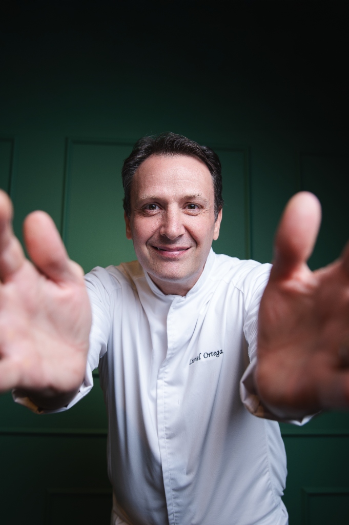 Chef francês Lionel Ortega de dólmã branco com as mãos estendidas para a câmera